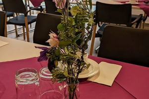 Vase mit vertrockneten Blumen auf einem mit rotem Tischtuch bedeckten Tisch in einem Restaurant mit gedeckten Tischen und schwarzen Stühlen im Hintergrund