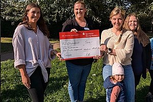 Vier Frauen stehen auf einer Wiese, eine hält einen großen roten Spendenscheck, ein Kleinkind mit Mütze steht davor