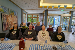 Mehrere ältere Menschen sitzen an einem Tisch mit blau-weiß karierten Tischdecken und bayerischen Herzanhängern in einem hellen Raum mit großen Fenstern und Bildern an der Wand.