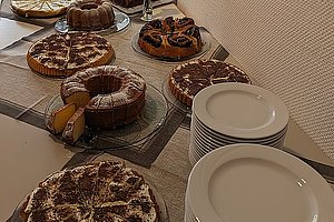 Mehrere verschiedene Kuchen auf einem gedeckten Tisch neben zwei Stapeln weißer Teller vor einer hellen Wand