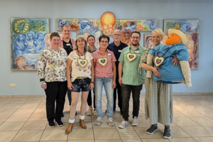 Gruppe von Menschen steht vor einer farbigen Wand mit mehreren Gemälden und einer großen Puppe in bunter Kleidung