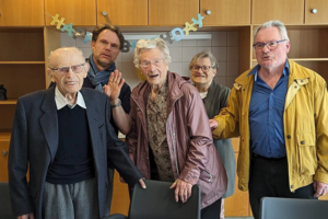 Fünf ältere Personen stehen in einer Küche vor einer Holzschrankwand mit einer Geburtstagsgirlande im Hintergrund.
