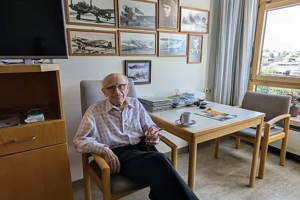 Mann sitzt in einem Stuhl in einem hellen Raum mit mehreren gerahmten Fotos und Urkunden an der Wand und einem Tisch mit Tassen und Zeitungen neben einem Fenster mit Blick nach draußen.