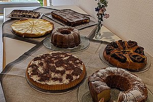 Mehrere verschiedene Kuchen und Torten auf Glastellern auf einem mit Leinentuch bedeckten Tisch vor einer hellen Wand