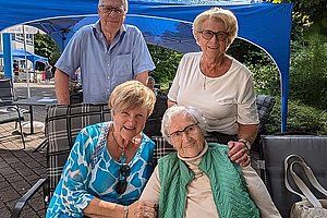 Vier ältere Personen sitzen und stehen vor einem blauen Pavillon im Freien, zwei sitzen auf Stühlen, zwei stehen dahinter, eine trägt eine grüne Weste