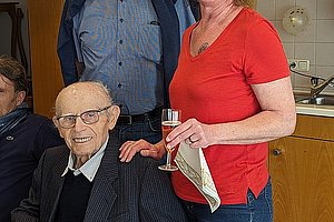 Drei Personen in einer Küche, eine Frau steht mit einem Glas in der Hand, zwei Männer sitzen und stehen daneben.