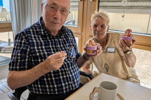 Zwei ältere Personen sitzen an einem Tisch, eine hält einen Löffel, die andere zeigt zwei Becher mit Eis oder Dessert