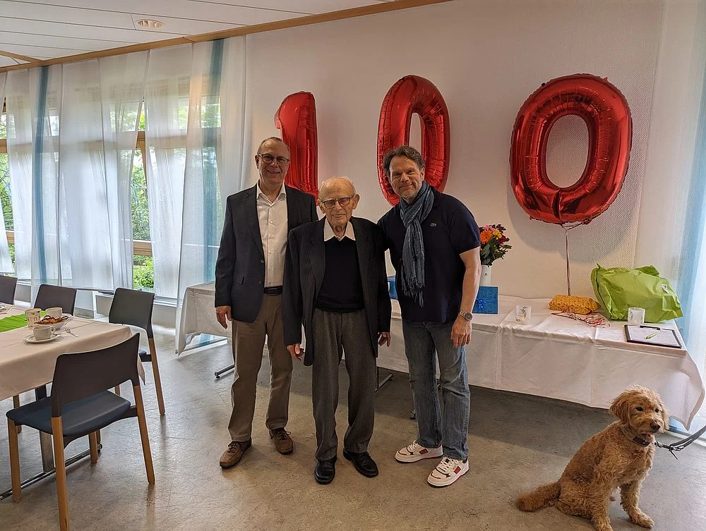 Drei Männer stehen nebeneinander in einem Raum mit großen Fenstern, im Hintergrund sind rote Luftballons in Form der Zahl 100 und ein sitzender Hund zu sehen.