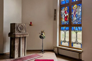 Innenraum einer Kapelle mit rotem Teppich mit grünem Kreuz, steinerner Kanzel und buntem Kirchenfenster