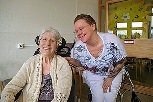 Ältere Frau im Rollstuhl mit hellbeiger Strickjacke neben einer tätowierten Pflegekraft in weißer Kleidung vor einer Tür mit Glasfenster und gelber Wanddekoration
