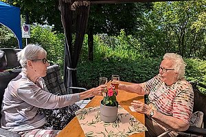 Zwei ältere Frauen sitzen an einem Holztisch im Freien und stoßen mit Gläsern an