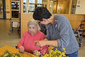 Zwei Frauen sitzen an einem Tisch mit gelben Blumen, eine steht und hilft der anderen beim Basteln oder Arrangieren der Blumen.