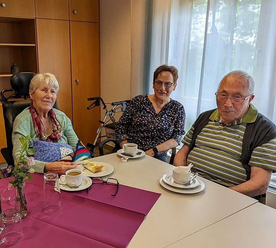 Drei ältere Personen sitzen an einem Tisch mit Tassen, Tellern und einem Stück Kuchen in einem hellen Raum mit Gardinen und einem Rollator im Hintergrund.