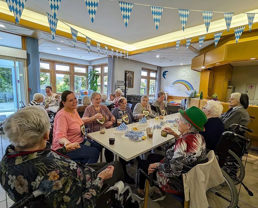 Gruppe älterer Menschen sitzt an einem langen Tisch in einem Gemeinschaftsraum mit blau-weißen Wimpelketten an der Decke