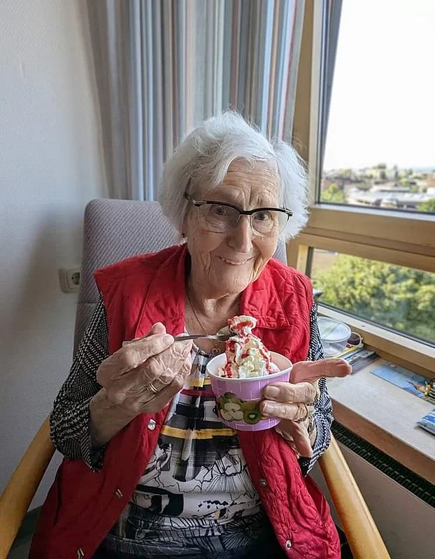 Ältere Person sitzt in einem Sessel am Fenster und hält eine Schale mit Eiscreme und Sahne in der Hand.