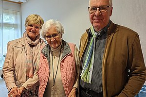 Drei Personen stehen in einem hellen Raum, zwei Frauen tragen leichte Jacken und Schals, ein Mann trägt eine braune Jacke und einen gestreiften Schal