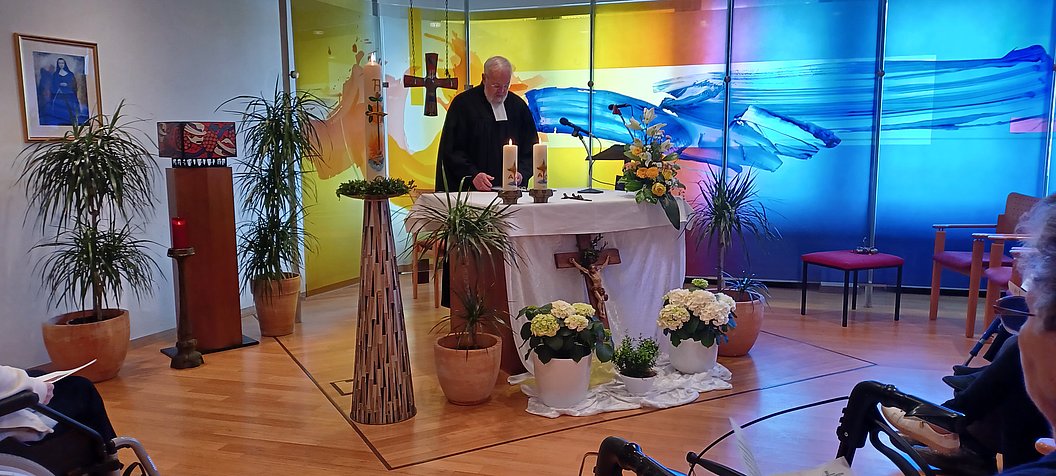 Ein Priester steht an einem Altar in einem hellen Raum mit bunten Glasfenstern, umgeben von Pflanzen und Blumen.