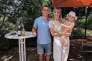 Drei Personen stehen im Freien auf gepflastertem Boden neben einem Stehtisch mit Getränken und einem Rucksack, eine Person hält ein Kind mit einem Hut und einen gelben Luftballon in der Hand, im Hintergrund Bäume und ein Sonnenschirm.