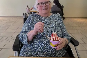 Person sitzt an einem Tisch mit einer Tasse Kaffee und hält eine Schale mit Eiscreme und roter Soße in der Hand, im Hintergrund hängen mehrere Bilder an der Wand.