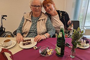 Zwei Personen sitzen an einem Tisch mit lila Tischdecke auf dem eine Tasse Kaffee, ein Teller mit Kuchen, eine Flasche Mineralwasser, eine kleine Schale mit Butter und ein Glas mit Blumen stehen