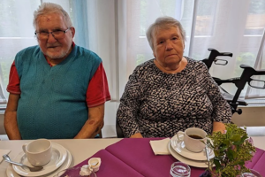 Zwei ältere Personen sitzen an einem mit lila Tischdecke gedeckten Tisch mit Kaffeetassen, Teller und einem kleinen Blumenarrangement