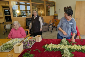 Drei Personen arrangieren in einem Raum mit Holzvertäfelung und Rollatoren auf einem roten Tuch mehrere Blumen zu einem großen Kreuz