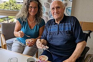 Zwei Personen sitzen an einem Tisch, eine hält einen Löffel mit Essen, auf dem Tisch stehen eine Tasse Kaffee, ein Glas Wasser und eine Schale mit Trauben.