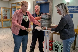 Drei Personen stehen um einen hohen Tisch mit einem Bierfass und einem Holzbrett in einem Raum mit blau-weißen Wimpeln an der Decke.