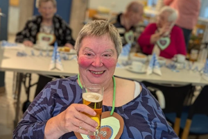 Person mit blau gemustertem Oberteil hält ein Glas und trägt ein Herz aus Lebkuchen mit Schriftzug um den Hals, im Hintergrund sitzen weitere Personen an Tischen.