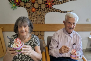 Zwei ältere Personen sitzen nebeneinander an einem Tisch und halten jeweils eine Schale mit Eis und Sahne in den Händen