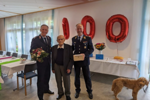 Zwei Männer in Uniform und ein älterer Mann stehen in einem Raum mit Tischen, im Hintergrund große Luftballons in Form der Zahl 100 und ein Hund mit Leine.