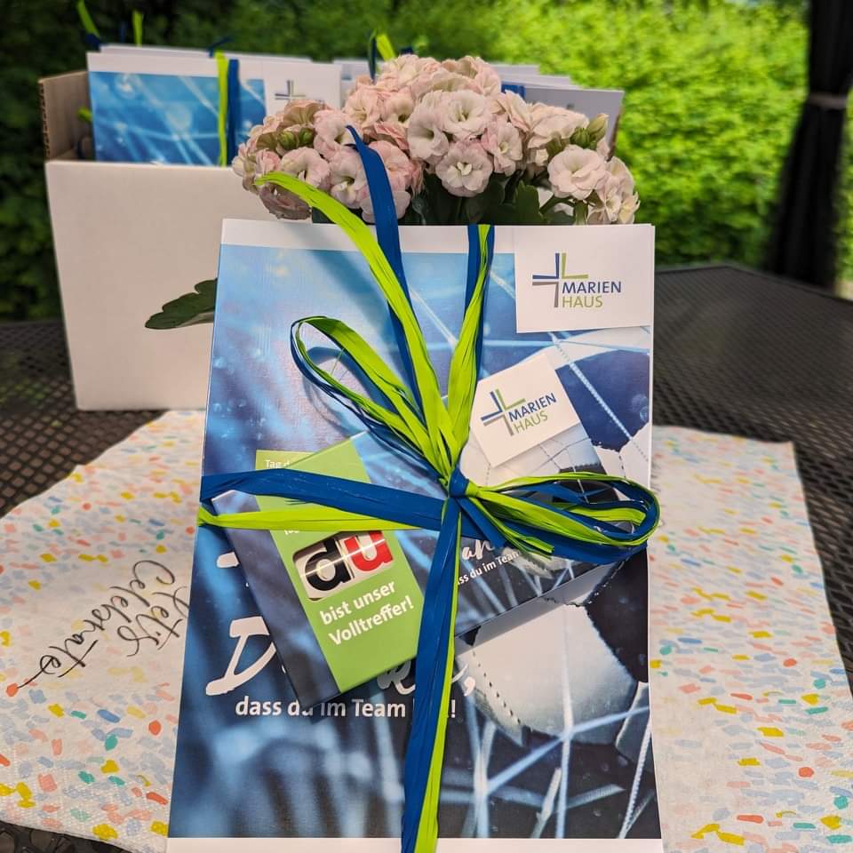 Geschenk mit blau-grünem Band, Karte mit Logo 'Marien Haus' und Text 'Du bist unser Volltreffer', auf Tisch mit bunter Tischdecke und Blumen im Hintergrund