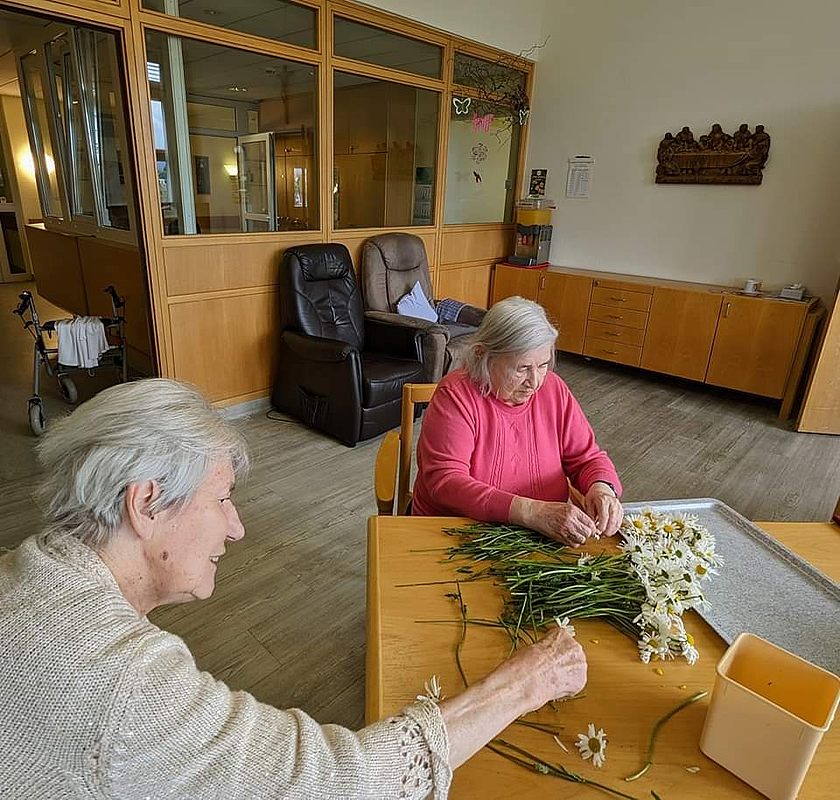 Zwei ältere Personen sitzen an einem Holztisch und binden Blumen zu Sträußen in einem Raum mit Holzverkleidung und Sesseln.