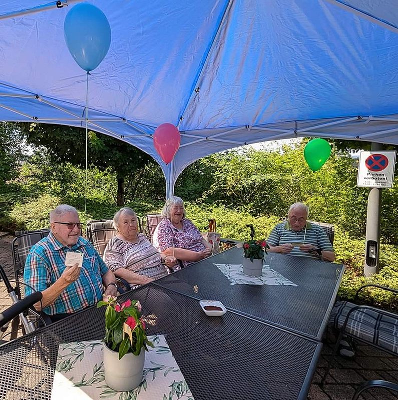 Vier Personen sitzen an einem rechteckigen Gartentisch unter einem Pavillon mit bunten Luftballons